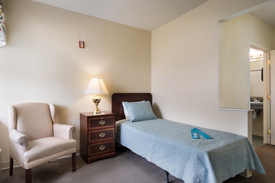A minimalist bedroom featuring a single bed, nightstand with a lamp, and an armchair.