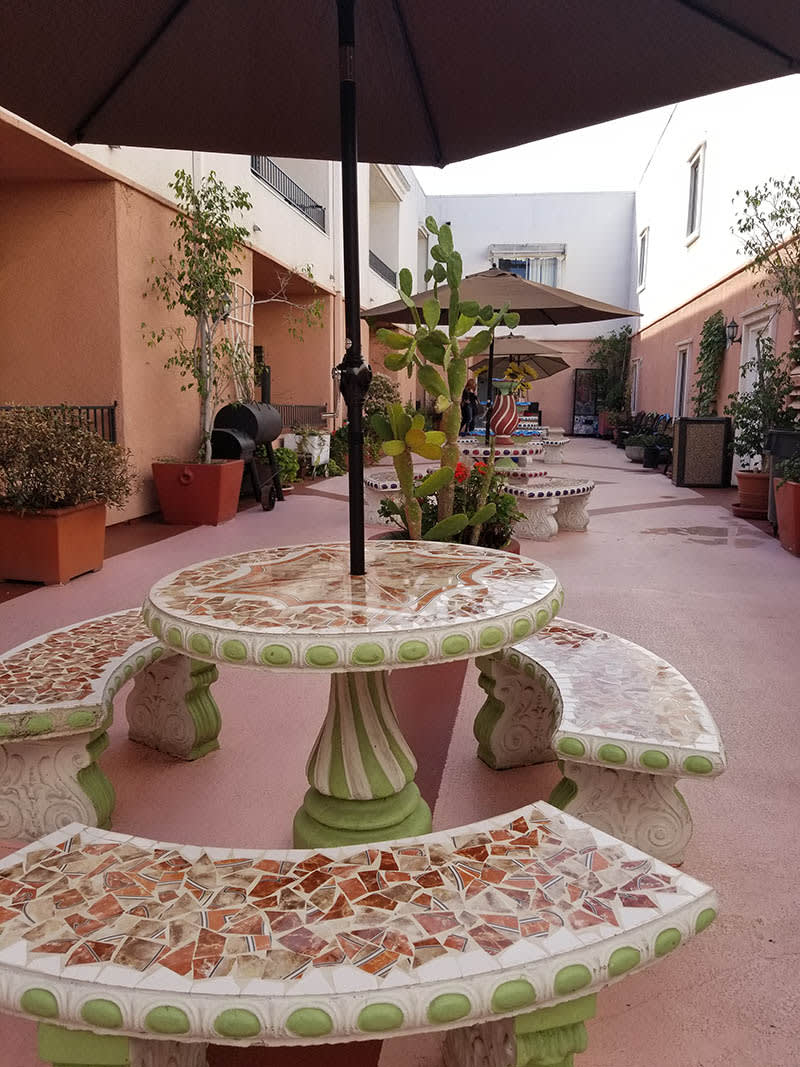 A cozy courtyard with mosaic tables and benches, surrounded by potted plants and shaded by umbrellas.