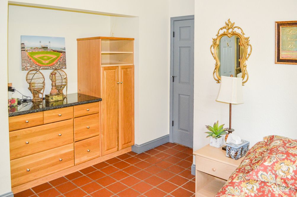 A cozy bedroom interior featuring a wooden dresser, a decorative mirror, and a bedside table with a lamp and plant.