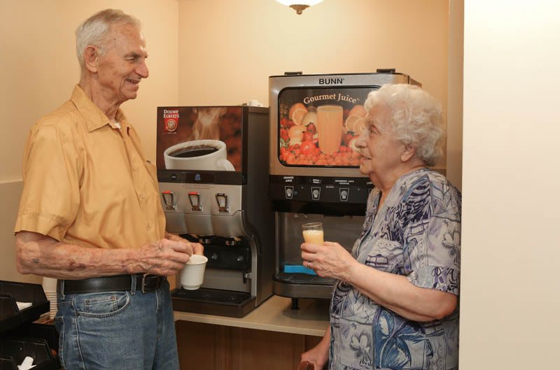 Two elderly individuals enjoy coffee and conversation by a beverage machine.
