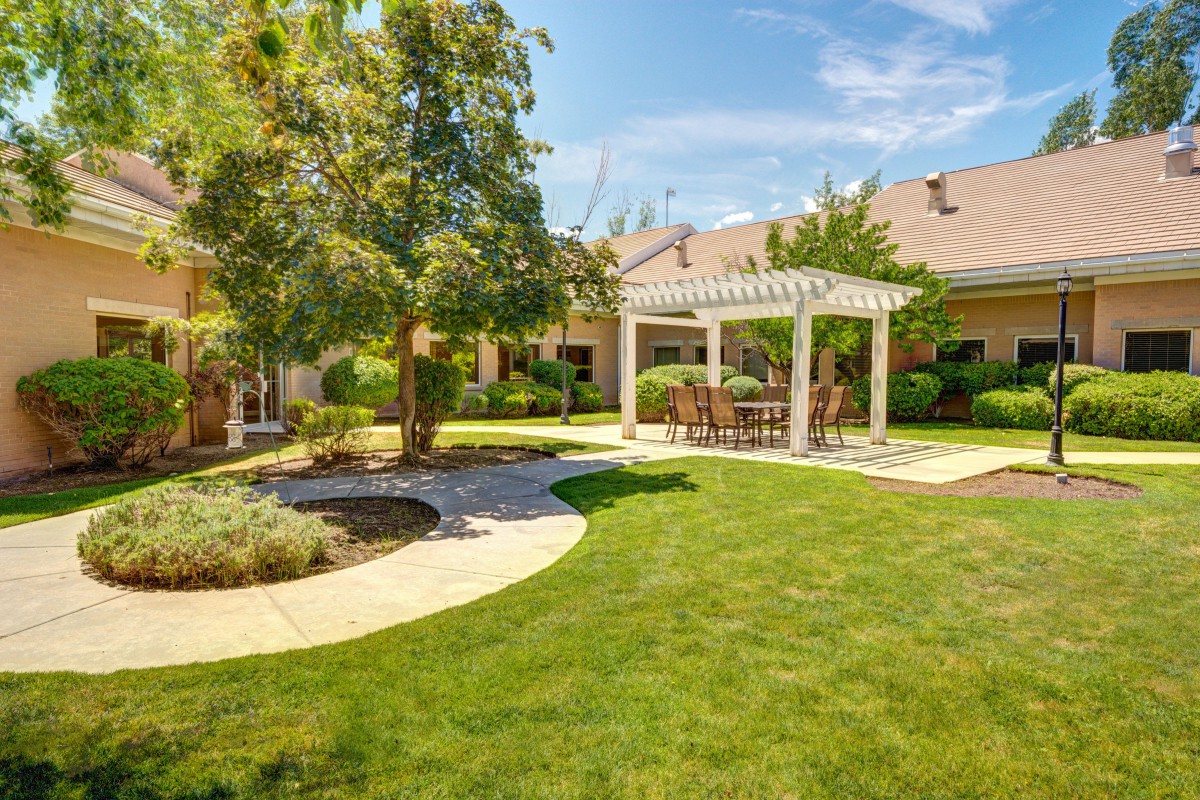 A beautifully landscaped backyard featuring a pergola with outdoor seating, surrounded by lush grass and trees on a sunny day.