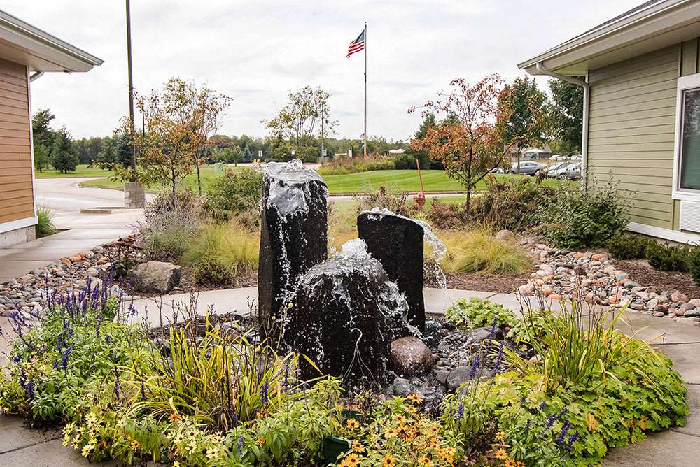 A tranquil garden scene featuring a stone fountain surrounded by lush greenery and an American flag in the background.