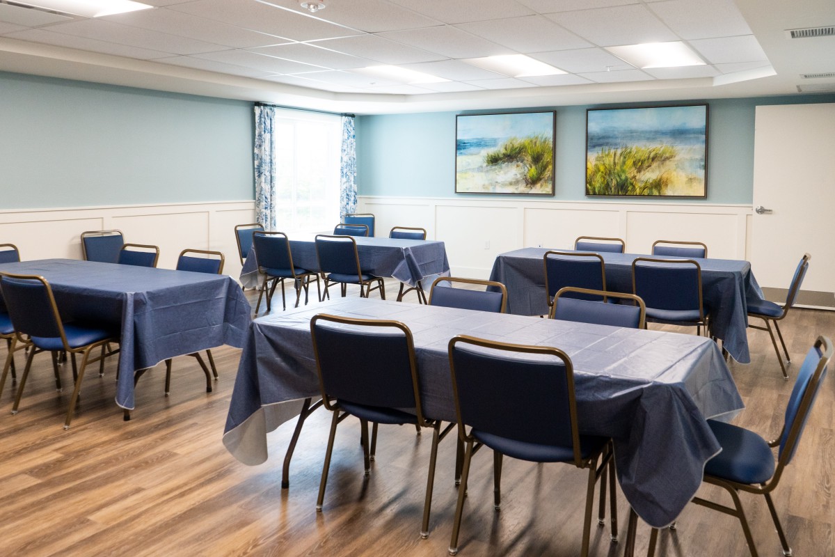 A neatly arranged meeting room with tables covered in blue tablecloths and chairs, featuring coastal artwork on the walls.