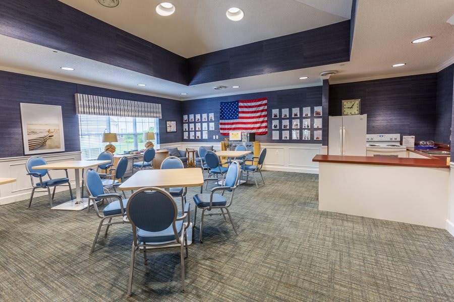 A well-lit meeting room featuring several tables and chairs, an American flag on the wall, and a row of windows.
