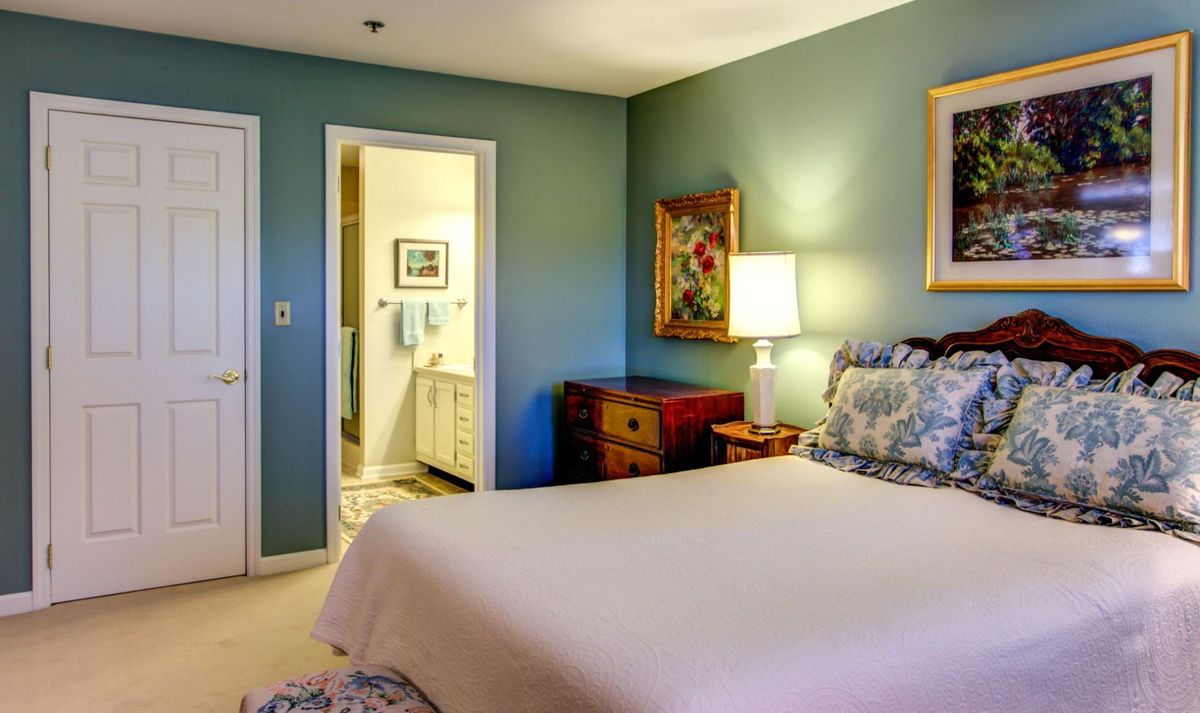A cozy bedroom with a neatly made bed, soft lighting from a lamp, and tasteful decor including paintings and furniture.
