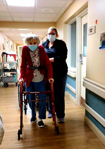 A caring healthcare worker assists an elderly woman using a walker in a hospital corridor.