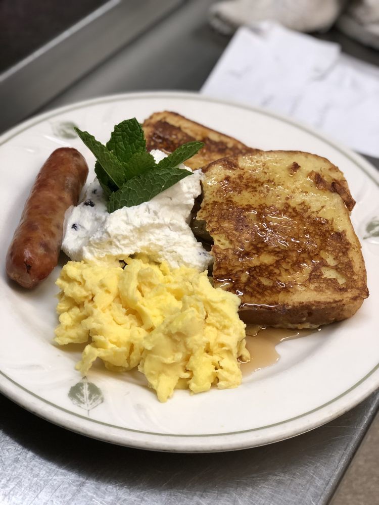 A plate of breakfast featuring scrambled eggs, a sausage, French toast topped with whipped cream and herbs.