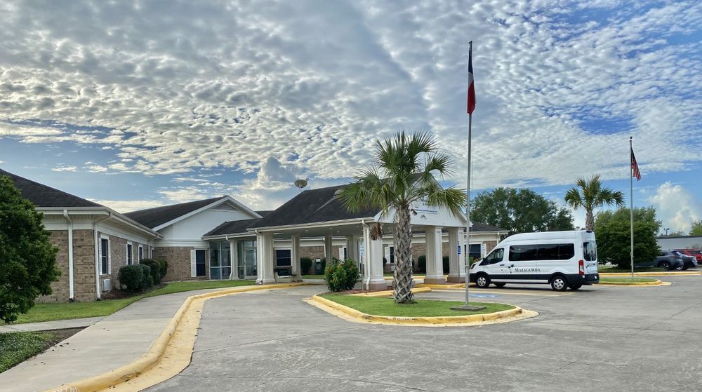 A building with a van parked in the driveway, featuring a flagpole and a palm tree under a cloudy sky.