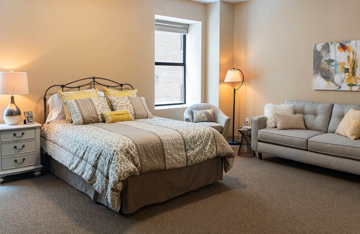 A cozy bedroom with a neatly made bed, a lamp, and a comfortable chair by the window.