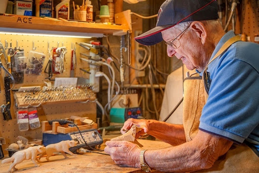An elderly man carefully works on a woodworking project in his well-equipped workshop.