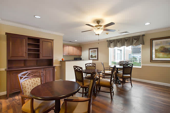 A cozy dining area with wooden tables, chairs, and a ceiling fan, adjacent to a small kitchenette and a large window with city views.