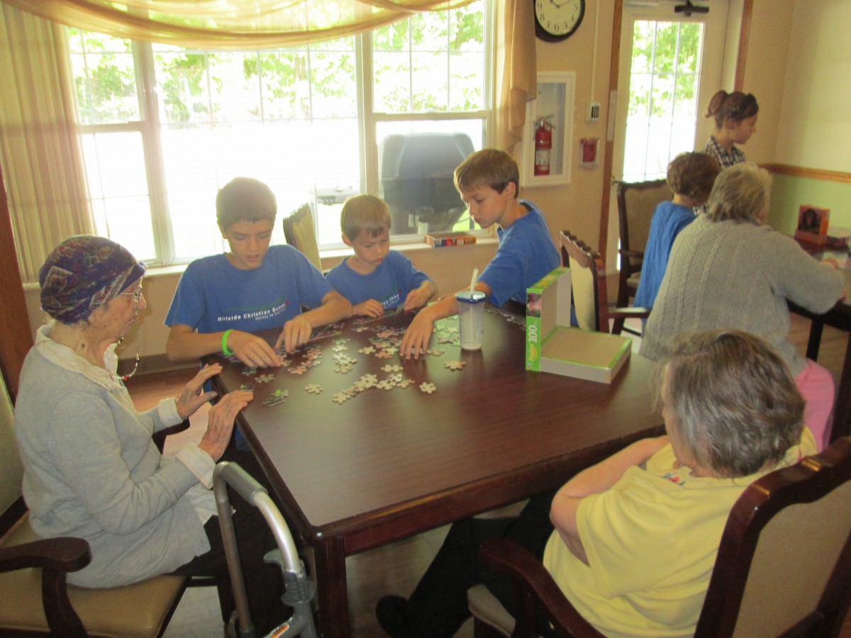 Children and elderly individuals working together on a puzzle at a table inside a room.