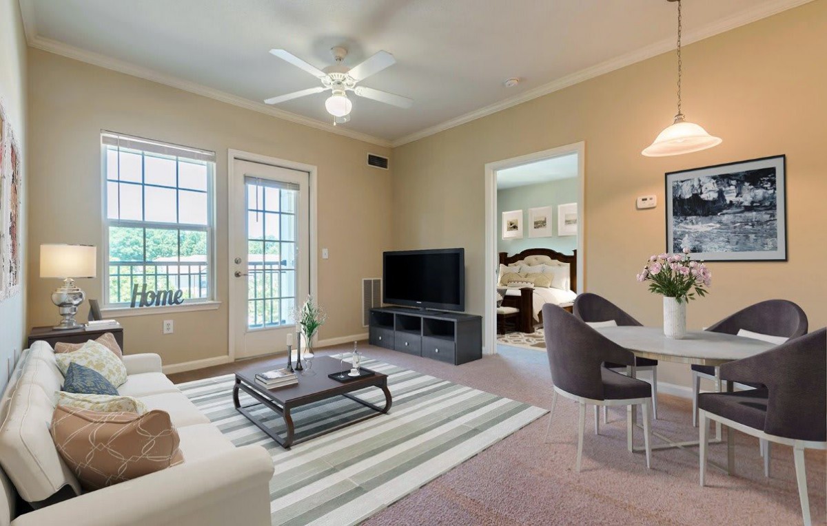A cozy living room with a TV, sofa, armchair, and coffee table, featuring neutral tones and natural light from large windows.
