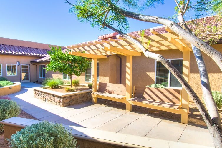 A sunny patio with a wooden pergola providing shade, surrounded by landscaping.