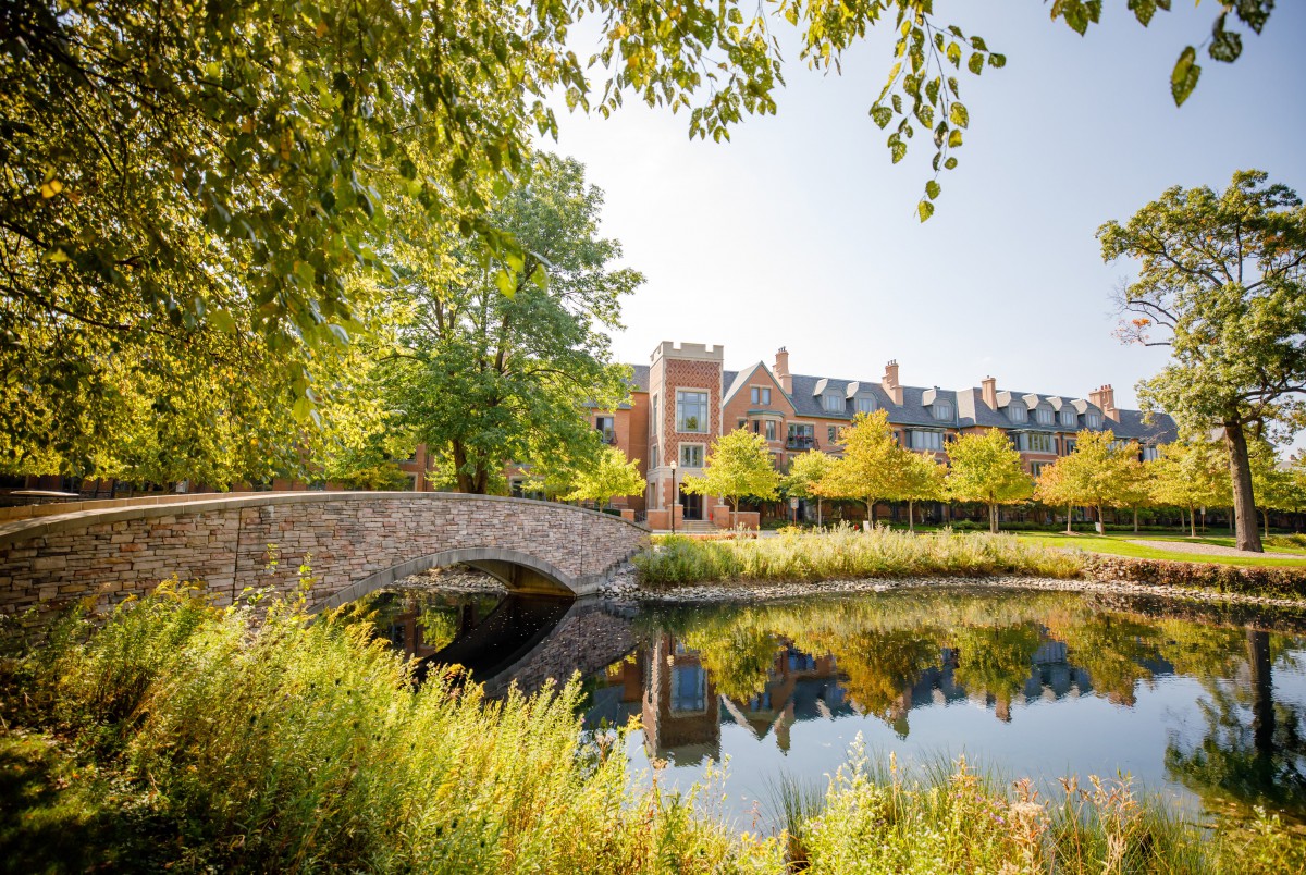 A scenic view of a stone bridge over calm water with a brick building and lush greenery in the background.