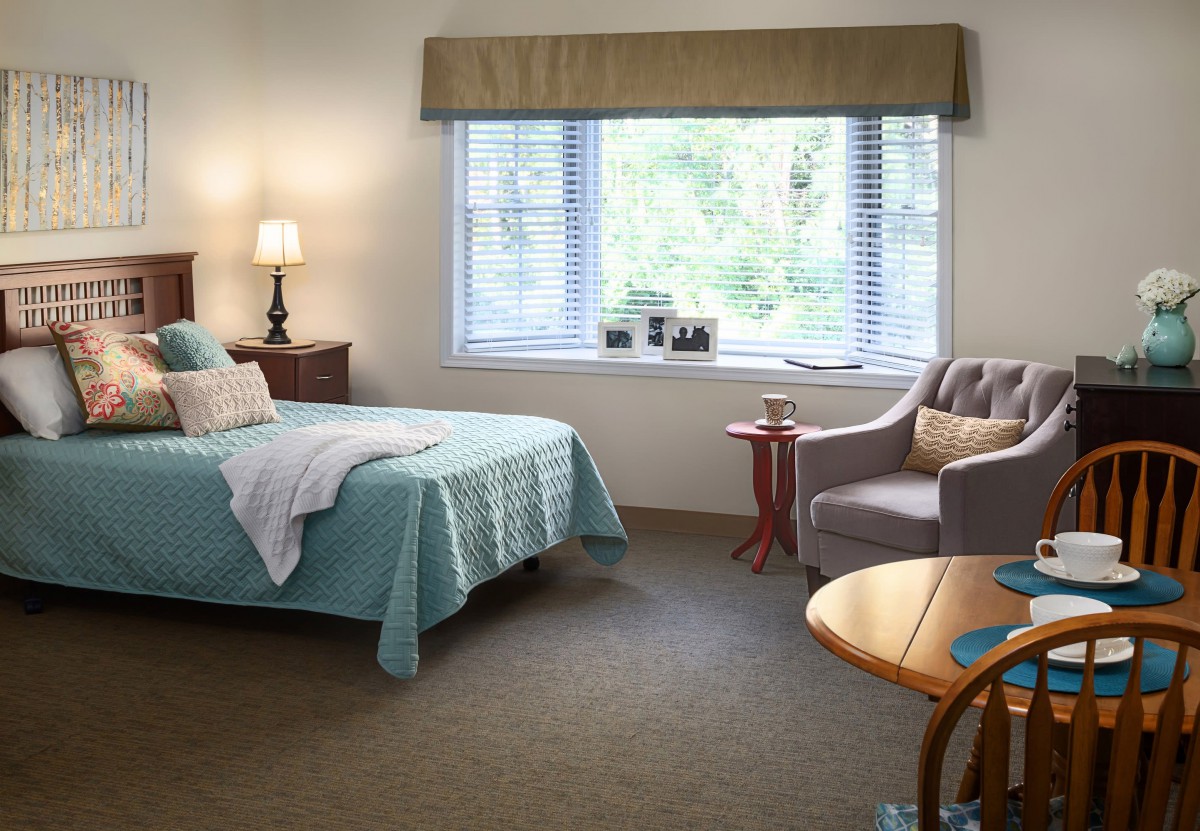 A cozy bedroom with a bed, lamp, and chair by a large window.