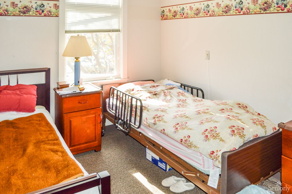 A cozy bedroom with a hospital bed next to a window, featuring a nightstand and a lamp.