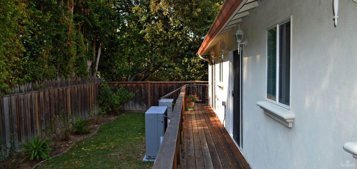 A wooden deck alongside a house with a yard and fence, illuminated by sunlight and surrounded by trees.