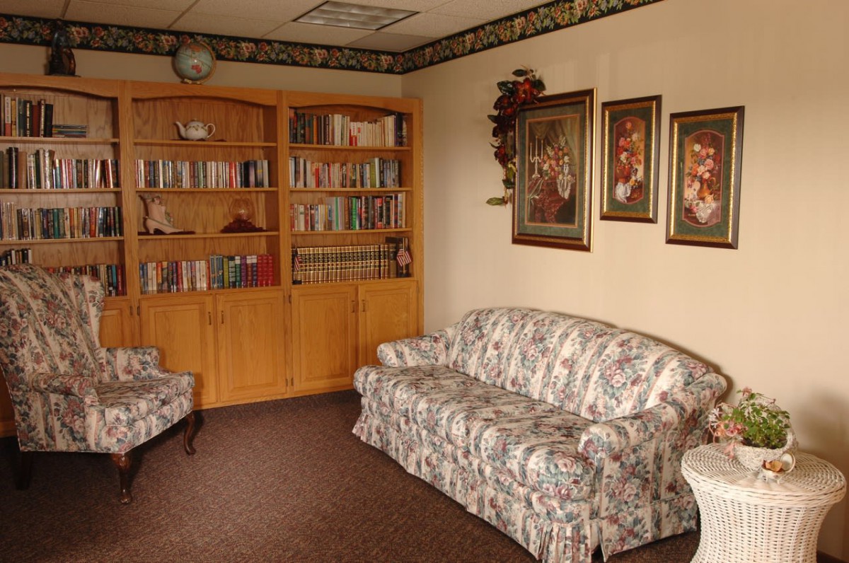 A cozy living room with a floral sofa, an armchair, a wooden bookshelf filled with books, and paintings on the wall.
