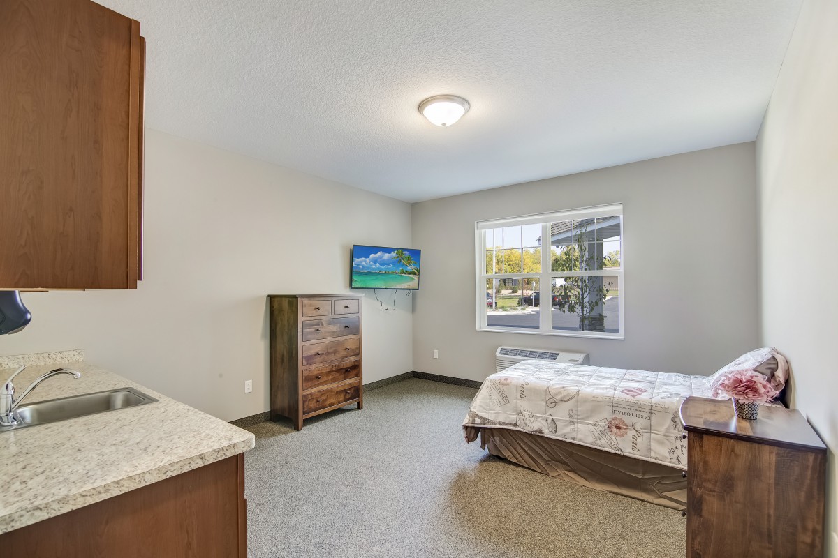 A minimalist bedroom featuring a bed, dresser, and wall-mounted TV with natural light coming through the window.