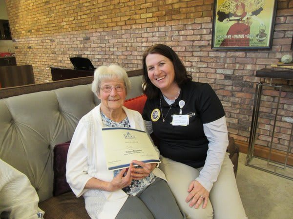 An elderly woman holding a certificate sits beside a nurse, both smiling at the camera.