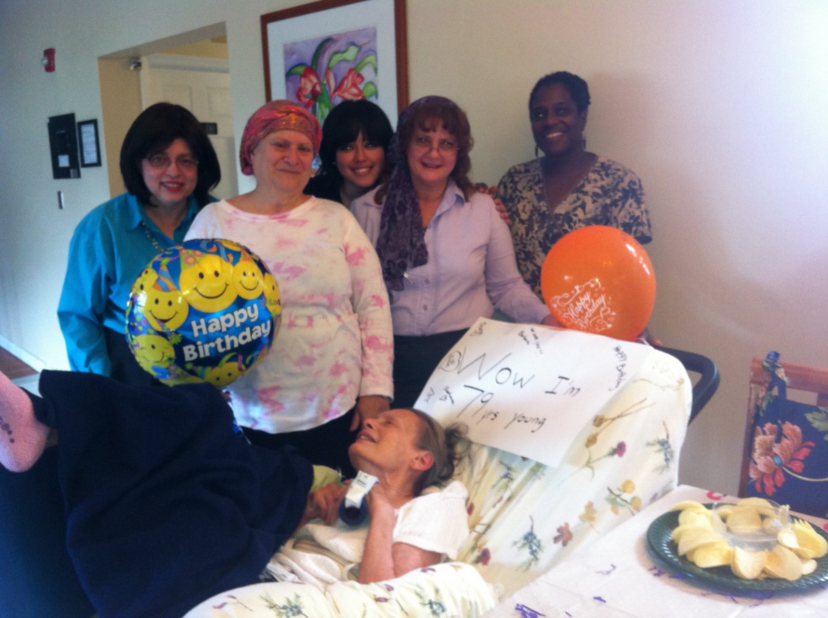 A group of people gathered around an elderly person in a hospital setting, holding balloons and smiling.