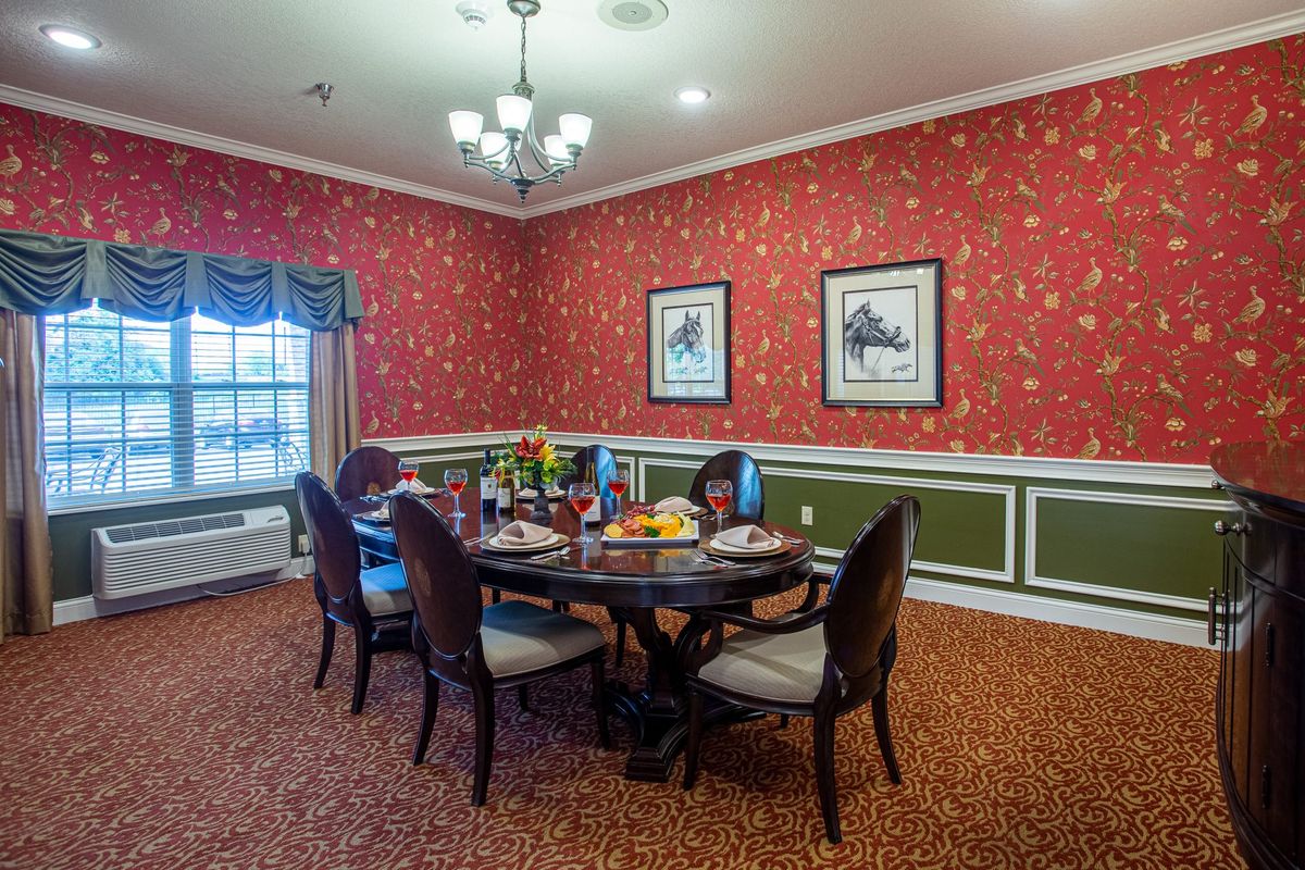 A traditional dining room with a round table, set for a meal, surrounded by elegant chairs and decorated with red wallpaper and framed artwork.