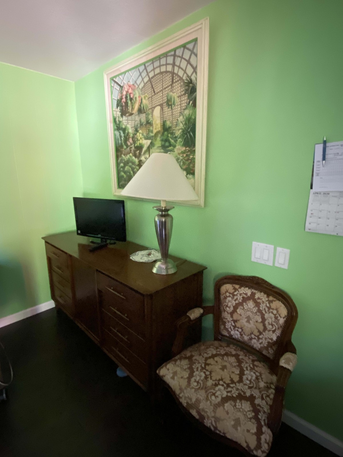 A cozy room with green walls featuring a wooden cabinet, a small television, an elegant vase, and an ornate chair beneath a large framed artwork.