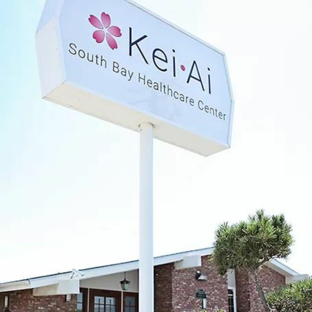 A sign for the Kei-Ai South Bay Healthcare Center outside a brick building.