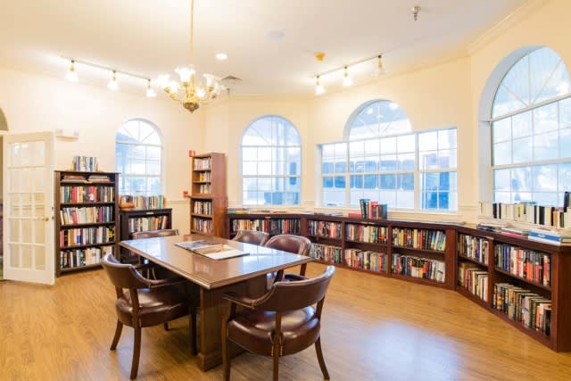 A cozy library with wooden shelves and a reading table, illuminated by natural light.