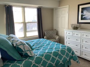 A cozy bedroom featuring a bed with blue patterned bedding, a white dresser, an armchair, and large windows with curtains.