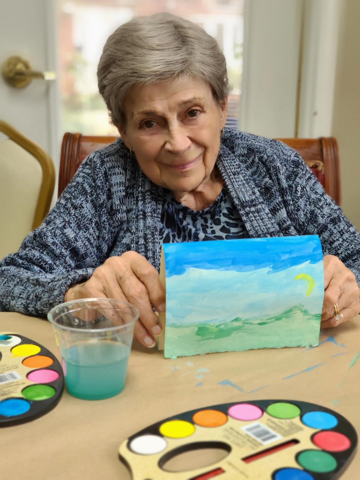 An elderly woman proudly displaying her watercolor painting indoors.