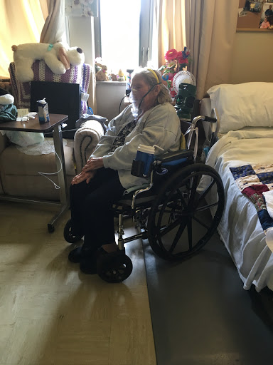 An elderly individual seated in a wheelchair within a hospital room, surrounded by personal belongings and care-related items.