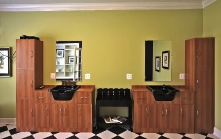 A modern barbershop interior featuring two wooden stations with black sinks and mirrors against a green wall, complemented by a black and white checkered floor.