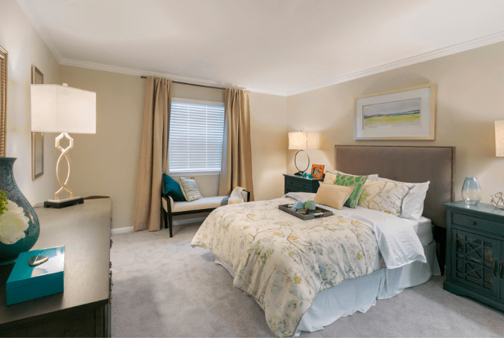 A cozy and well-decorated bedroom with a neatly made bed, bedside lamp, armchair near the window, and soft lighting.