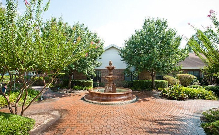 A tranquil garden setting featuring a central fountain surrounded by lush trees and a brick-paved walkway.