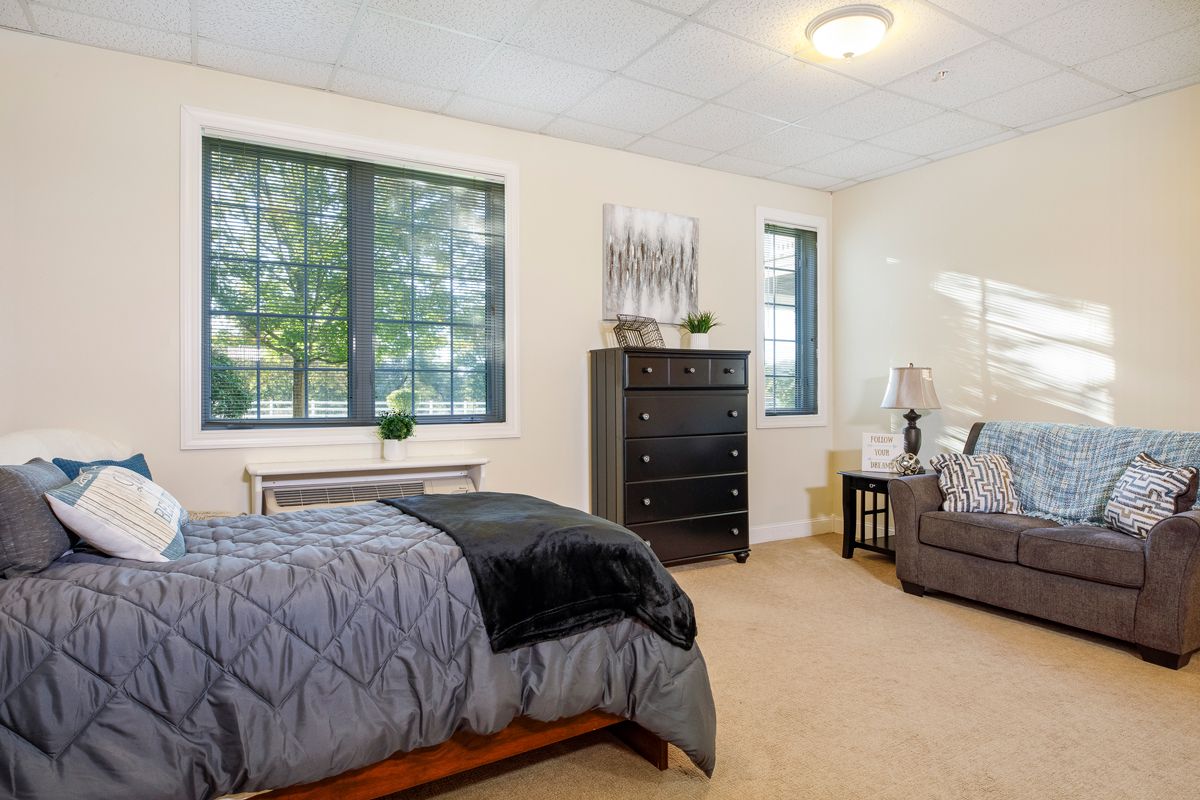 A cozy bedroom featuring a bed, dresser, armchair, and large windows.