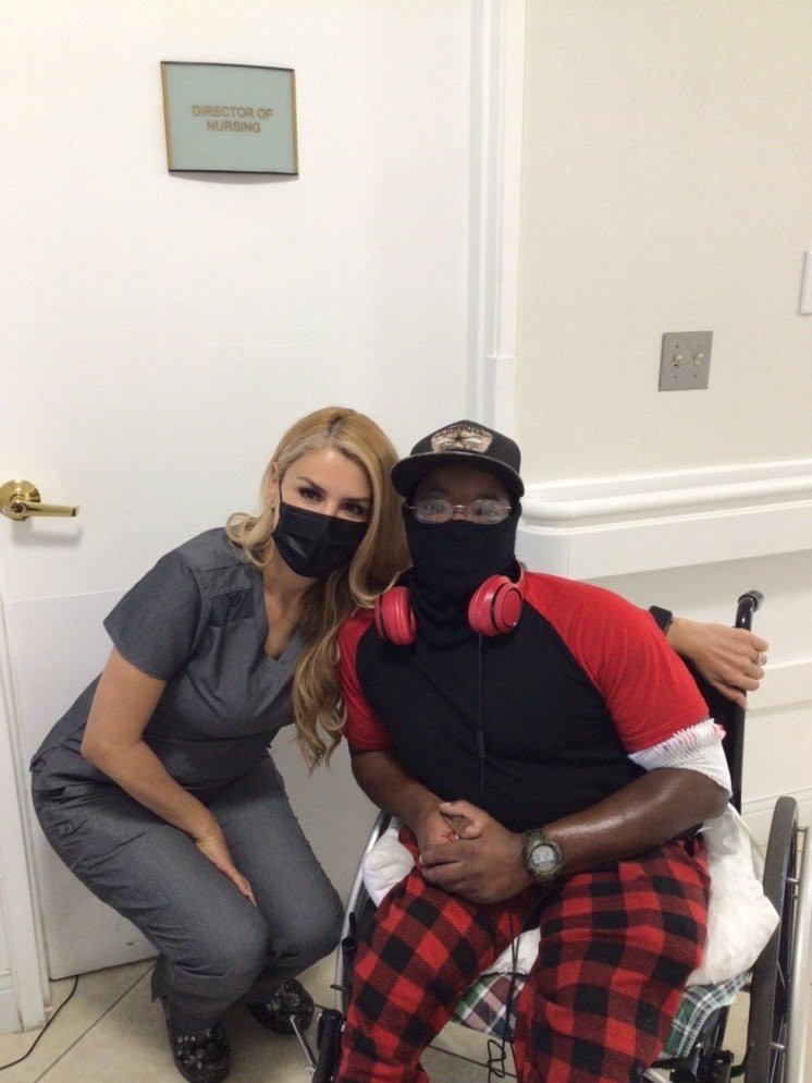 Two people in a healthcare setting wearing masks, one person is seated in a wheelchair.