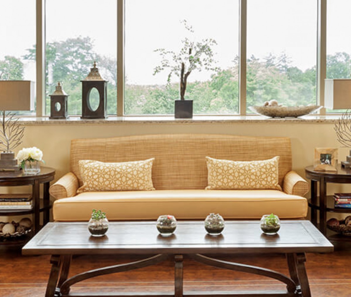 A cozy living room featuring a beige sofa with decorative pillows, complemented by a wooden coffee table and indoor plants near large windows.