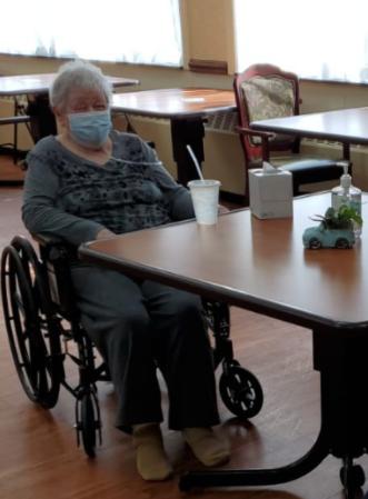 An elderly woman in a wheelchair, wearing a mask, seated at a table in a care facility.