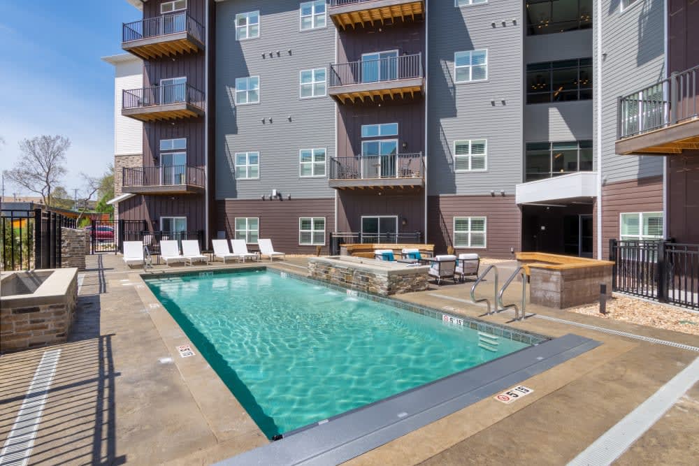 An outdoor swimming pool surrounded by lounge chairs in front of a modern apartment building with multiple balconies.