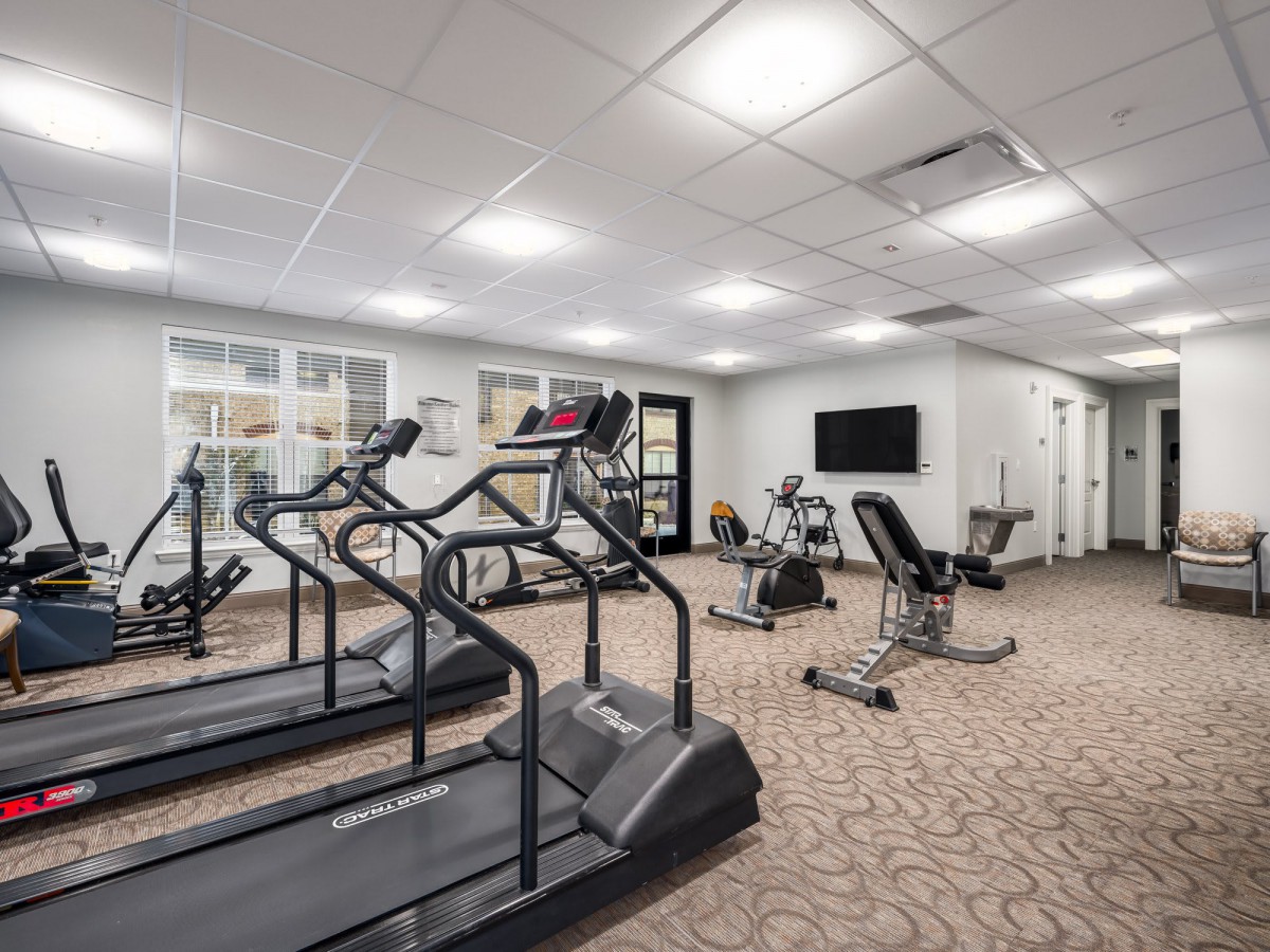 A well-equipped gym with treadmills, an exercise bike, and a weight bench under bright overhead lighting.