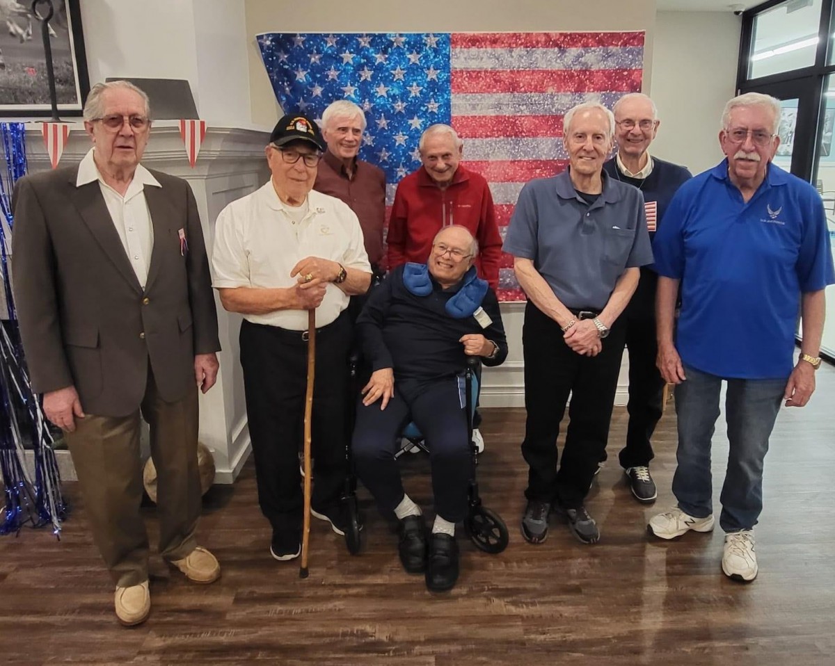 A group of elderly veterans posing together in front of an American flag indoors.
