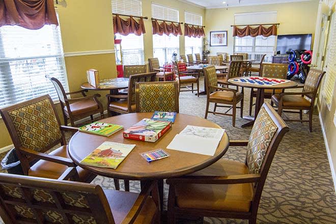 A cozy room with several tables and chairs, featuring books on the tables and large windows allowing natural light.