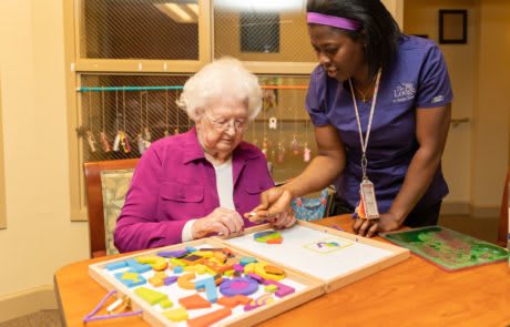 An elderly woman is engaged in a puzzle activity with the assistance of a caregiver.