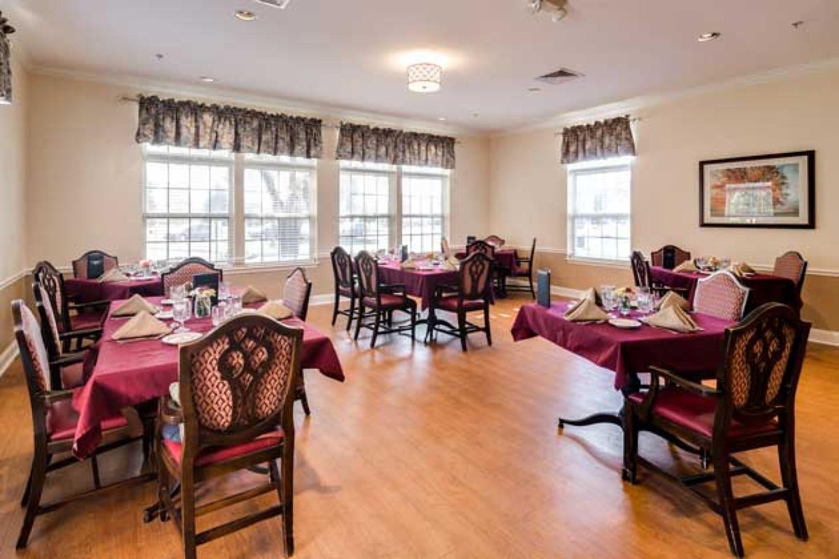 A cozy dining area with elegantly set tables and chairs, featuring large windows and wooden flooring.