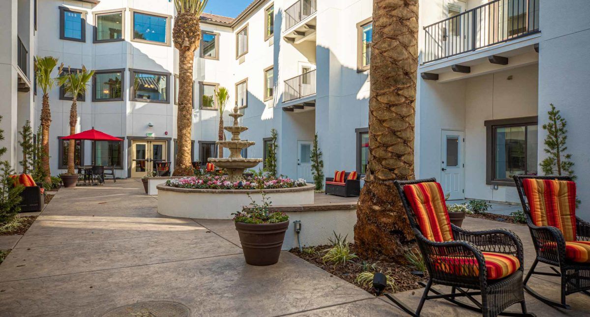 A sunny courtyard featuring palm trees, a fountain, and outdoor seating in a building complex.