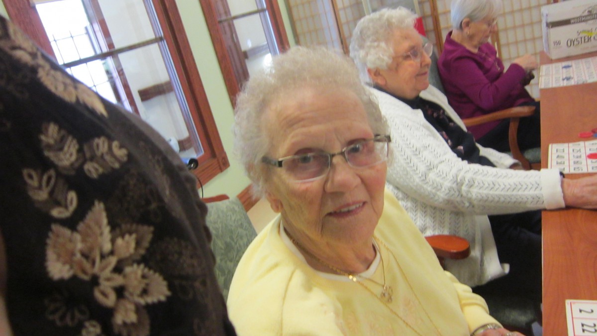An elderly woman with glasses seated indoors, smiling at the camera.
