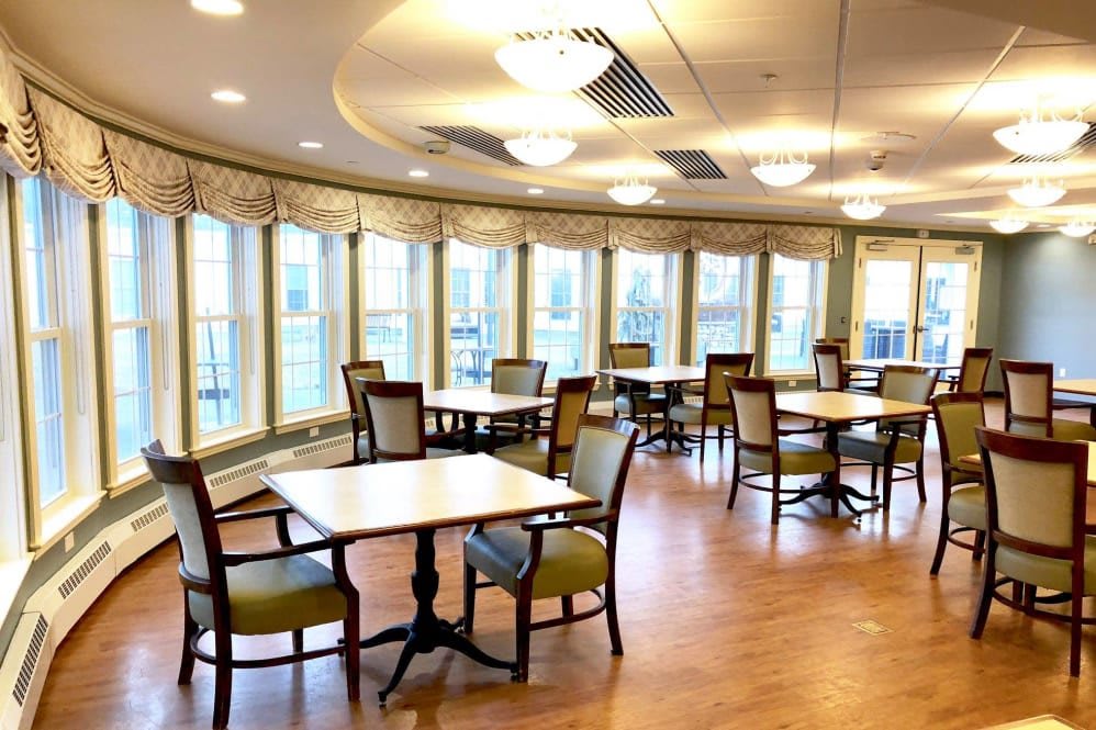 A cozy dining room with tables and chairs, featuring large windows and warm lighting.