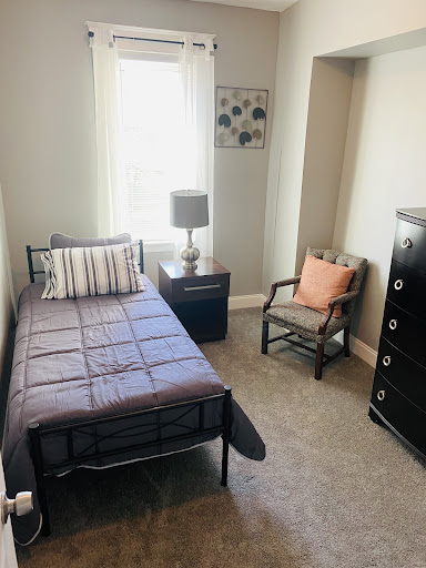 A cozy and minimalistic bedroom featuring a single bed, a nightstand with a lamp, an armchair, and a dresser.